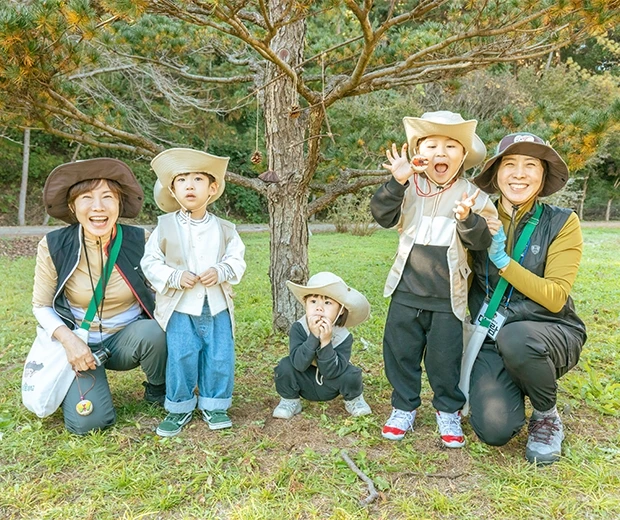 소나무 아래 모자를 쓴 아이들과 숲 해설사 선생님들이 나란히 앉아 함께 기념사진을 찍는 모습