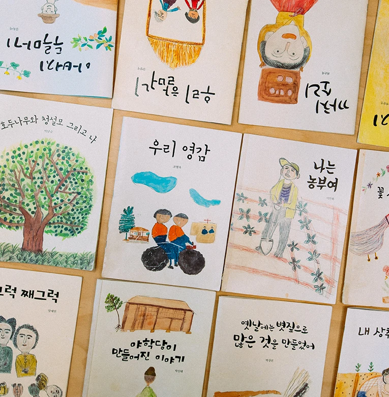 마을 어르신들이 직접 쓰고 그린 그림책들이 나란히 펼쳐진 모습, 서툴지만 정겨운 그림과 손글씨가 가득하다