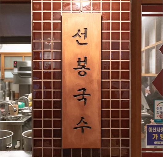 갈색 타일 벽에 걸린 선봉국수 구리 간판과 그 옆 주방 모습