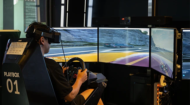 세 개의 화면을 보며 VR 기기를 쓰고 운전대를 잡은 레이싱 어트랙션 체험자