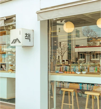 책 글자 간판이 달린 서점 모퉁이, 통유리 너머로 원목 의자와 책장이 보이는 거리 풍경
