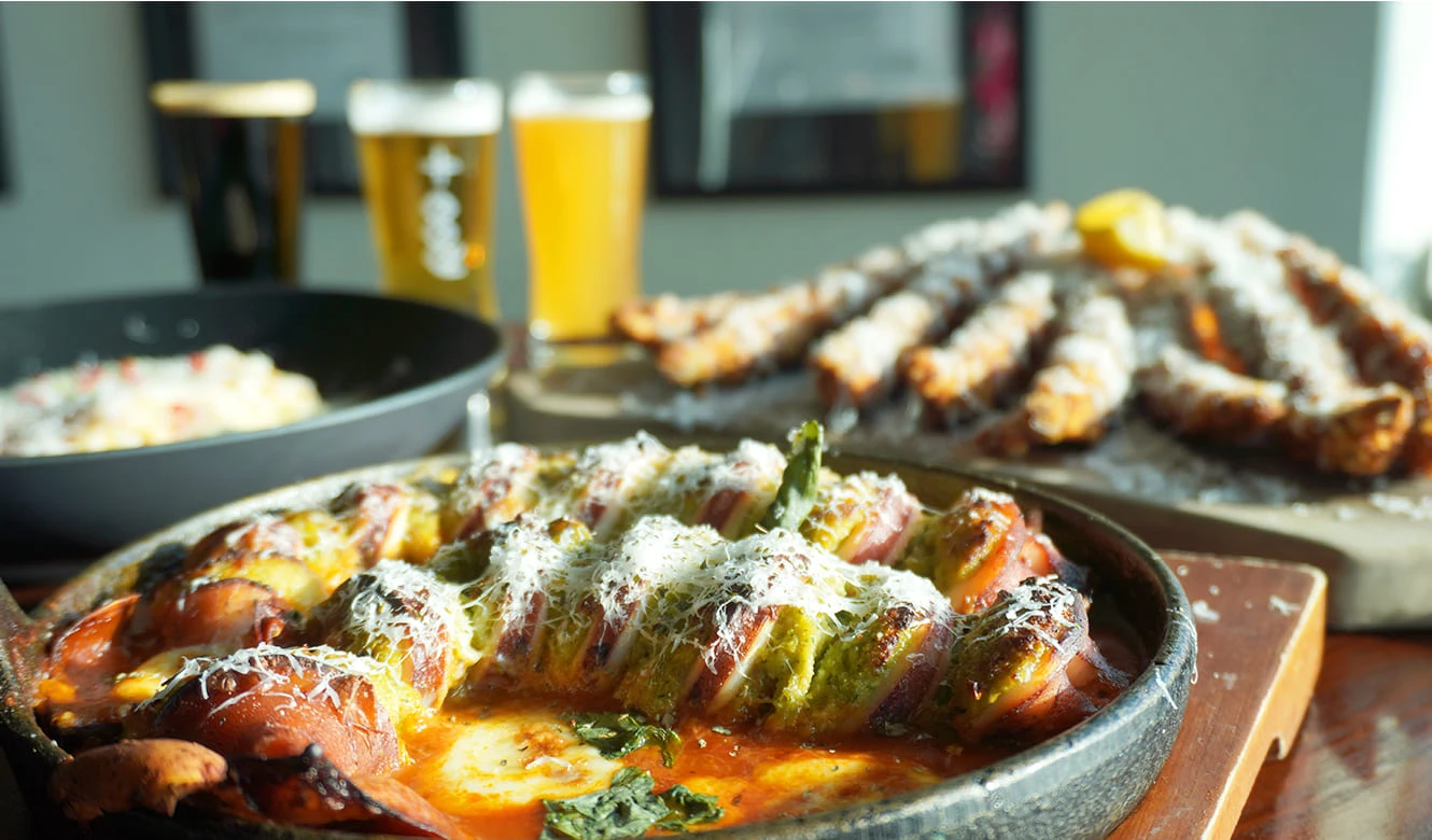 검은 무쇠 팬에 치즈를 얹어 구운 요리와 맥주, 빵 꼬치가 함께 차려진 크래프트루트의 겨울 특선 한 상