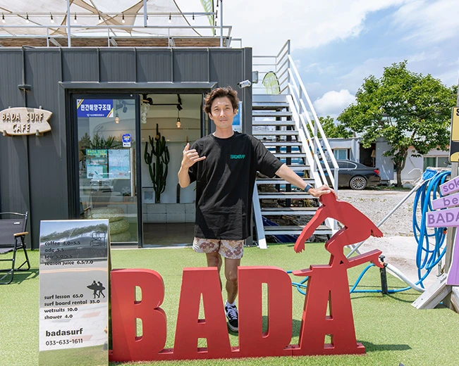 바다 서프 카페 앞에서 한 남성이 붉은 BADA 조형물에 손을 얹고 서핑 손짓을 하며 서 있다