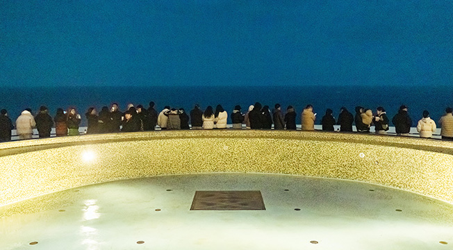 곡선형 수영장 난간에 기댄 사람들이 아직 어둑한 새벽 바다를 바라보며 일출을 기다린다