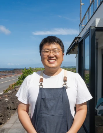 멜빵 앞치마를 두른 토끼썸 김병호 대표가 바다를 등지고 환하게 미소 짓고 있다