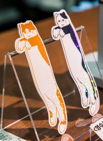 투명 거치대에 세워 둔 길쭉한 고양이 모양 일러스트 책갈피 두 개
