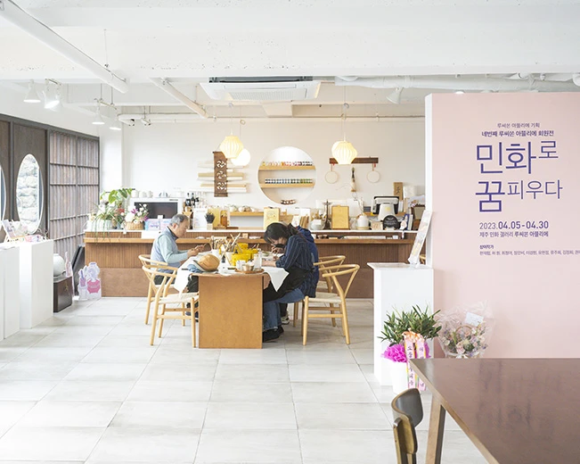 한가로운 공방 안에서 두 사람이 테이블에 앉아 작업하는 루씨손아뜰리에 내부, 전시 안내판이 세워진 모습