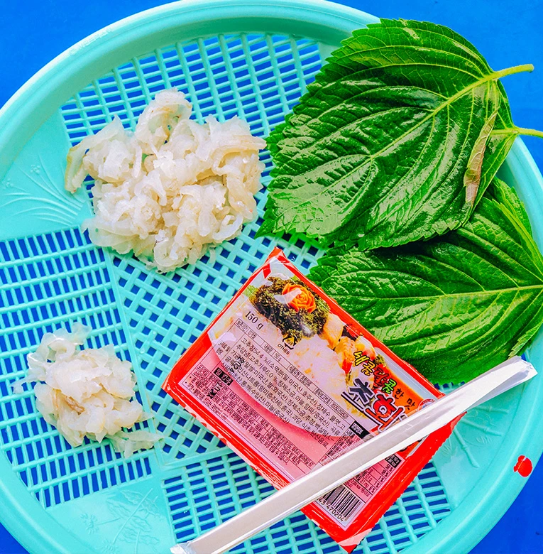 파란 소쿠리에 흰 살 회와 깻잎을 담고 초장 한 봉을 곁들인 모습