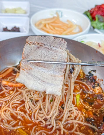 젓가락으로 집어 올린 수육 한 점과 그 아래 붉은 육수에 담긴 물막국수 클로즈업