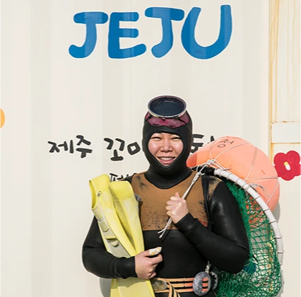 JEJU 글자가 적힌 벽 앞에서 테왁과 오리발을 들고 환하게 웃는 잠수복 차림 해녀
