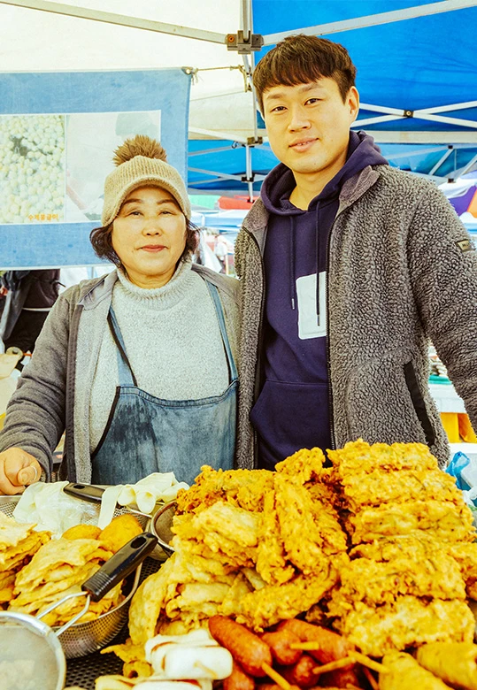 어묵 튀김 좌판 앞에서 나이 든 여성 상인과 젊은 남성이 나란히 서서 카메라를 보고 있고, 앞에는 갓 튀긴 어묵이 쌓여 있다