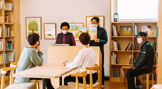 마스크를 쓴 어르신이 방문객들에게 직접 그린 그림책을 읽어주는 모습, 책장과 액자로 둘러싸인 아늑한 공간