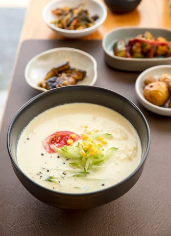 사계반상의 한식 한 상. 짙은 색 그릇에 담긴 고소한 크림빛 냉국 위에 방울토마토와 오이채, 옥수수가 올려져 있고 주변으로 여러 가지 밑반찬이 정갈하게 놓여 있다.
