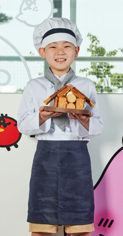 흰 요리사 모자와 앞치마 차림의 아이가 직접 만든 과자집을 들고 환하게 웃는 모습