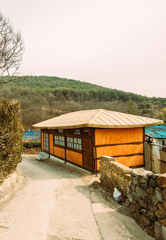 주황빛 흙벽과 초가지붕을 인 야학당 건물, 돌담과 산을 배경으로 마을 길가에 자리한 모습