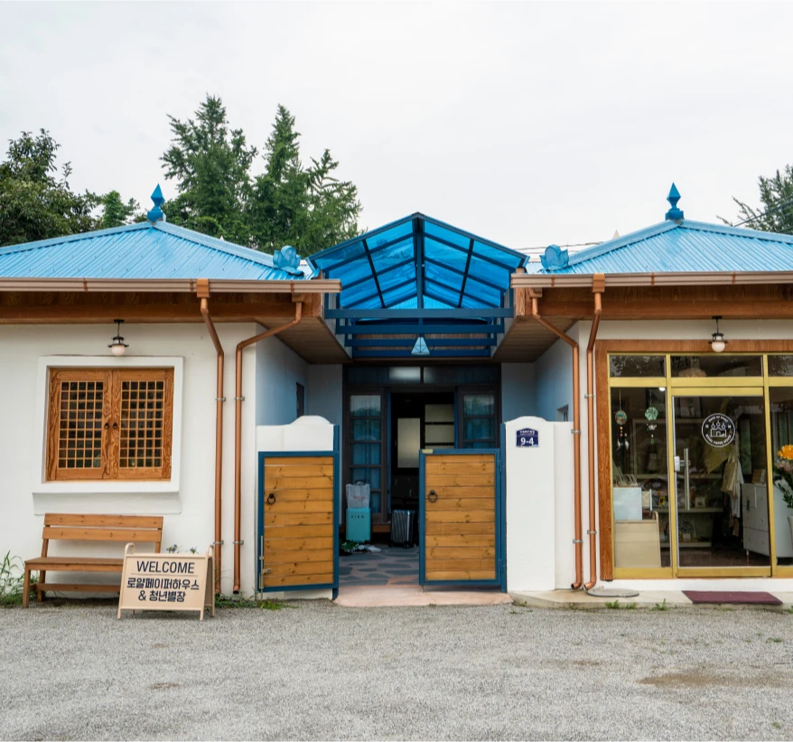 파란 지붕과 흰 벽이 어우러진 로얄페이퍼하우스 건물 외관, 입구에 환영 안내판이 놓인 모습