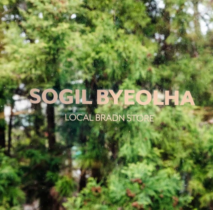 통유리창에 새겨진 SOGIL BYEOLHA LOCAL BRAND STORE 사인 너머로 짙푸른 숲이 비치는 스토어 입구