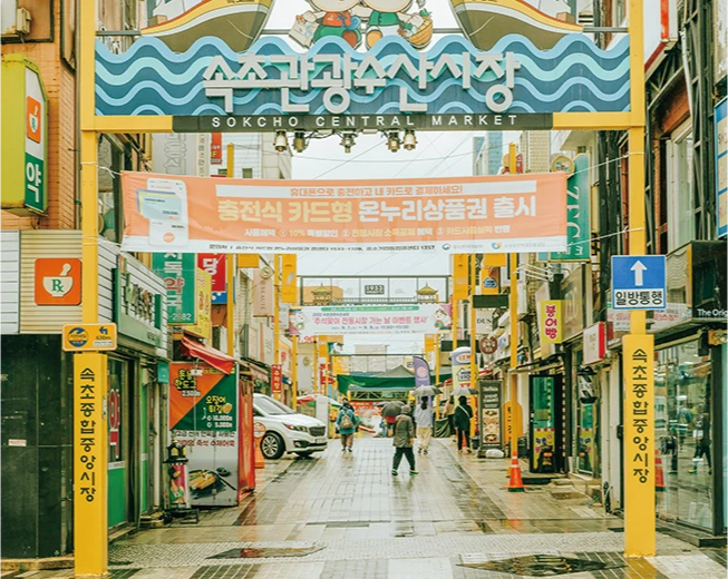 속초관광수산시장 입구의 화려한 아치 간판과 상품권 출시 현수막, 사람들이 오가는 거리 풍경