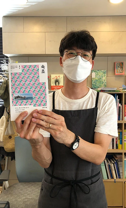 앞치마를 두른 마스크 쓴 남성이 속초를 소재로 한 책을 두 손으로 들어 보이는 모습