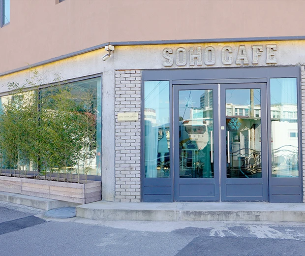 벽돌과 통유리로 된 모퉁이 건물에 SOHO CAFE 간판이 걸린 카페 외관