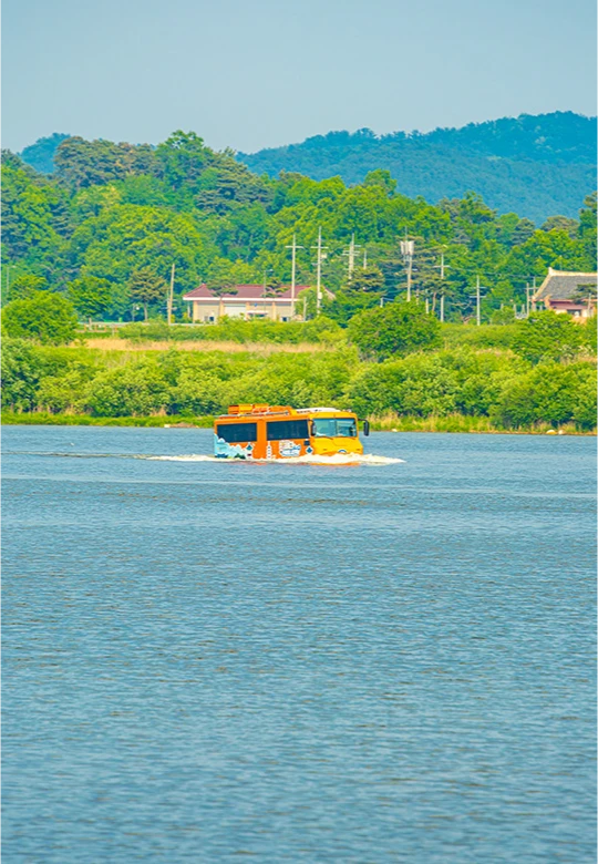 산과 마을을 배경으로 잔잔한 강 위를 나아가는 주황색 수륙양용버스의 모습