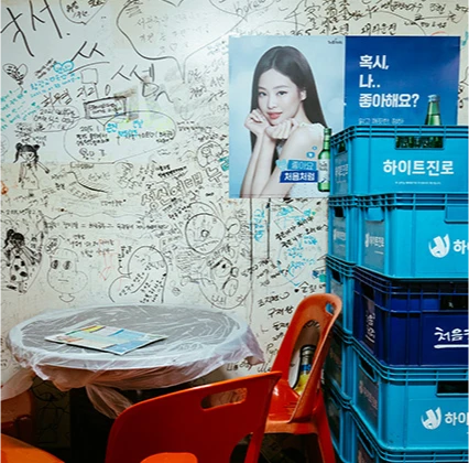 손님들의 낙서로 가득한 벽 옆에 주류 상자가 쌓이고 주황색 의자가 놓인 포장마차 내부
