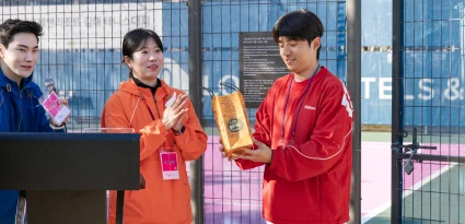 주황색 운동복의 진행 요원들이 박수치는 가운데 빨간 옷의 참가자가 경품을 받는 장면