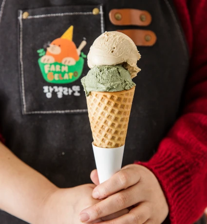 검정 앞치마 차림의 대표가 갈색과 녹색 두 가지 젤라또를 얹은 와플콘을 두 손으로 감싸 든 모습