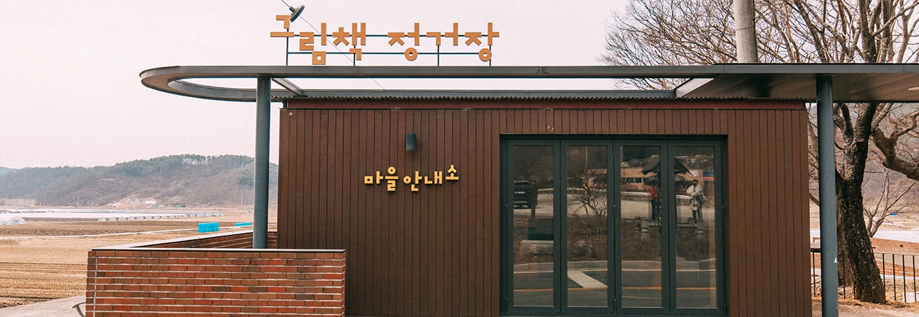 물가에 자리한 그림책 정거장, 나무로 지은 작은 버스정류장 건물 위에 새 조형물이 놓인 모습