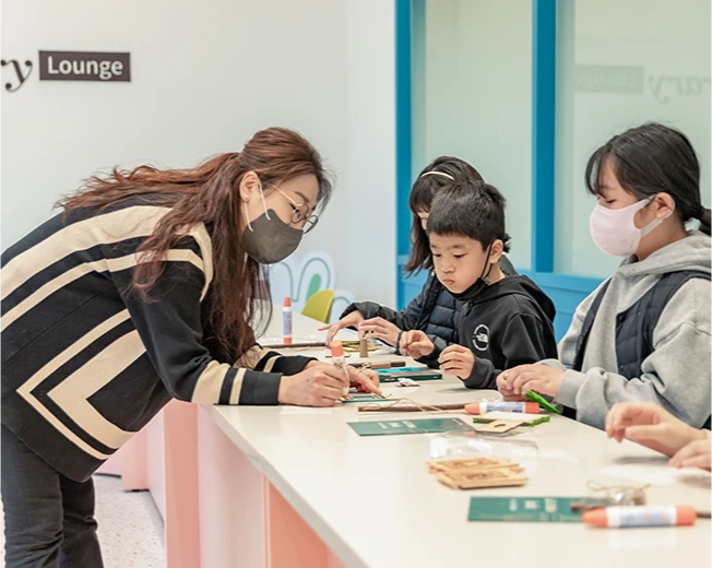 강사가 책상 너머로 몸을 숙여 아이들의 만들기 체험을 도와주는 모습