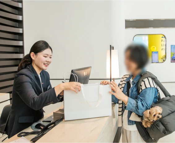 프런트 직원이 반려견을 안고 온 투숙객에게 펫 웰컴 기프트가 담긴 쇼핑백을 건네는 장면