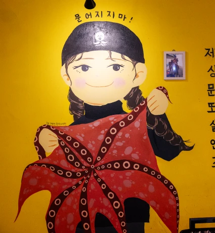 노란 벽에 그려진 문어를 두 손으로 펼쳐 든 두건 쓴 소녀 캐릭터 벽화, 위에 문어지지마 문구가 적힌 모습