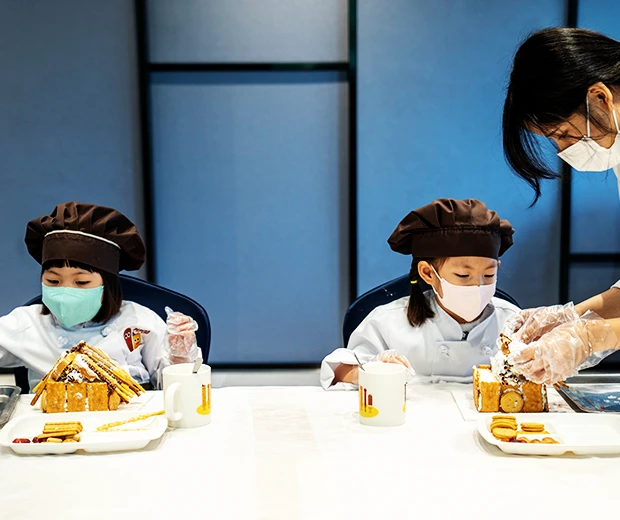 두 아이가 나란히 앉아 각자 과자집을 만들고 강사가 옆에서 손을 거들어 주고 있다