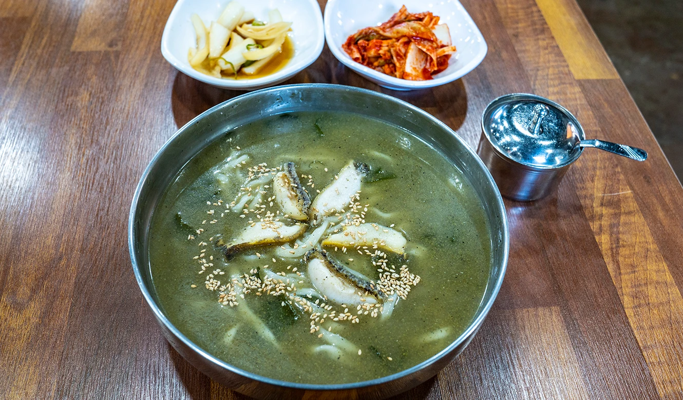 스테인리스 그릇에 초록빛 국물과 전복살을 담고 깨를 뿌린 전복칼국수, 곁에 김치 반찬