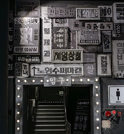 옛 간판이 빼곡히 붙은 흑백 벽면과 전구로 둘러싼 노형수퍼마켙 지하 입구 계단