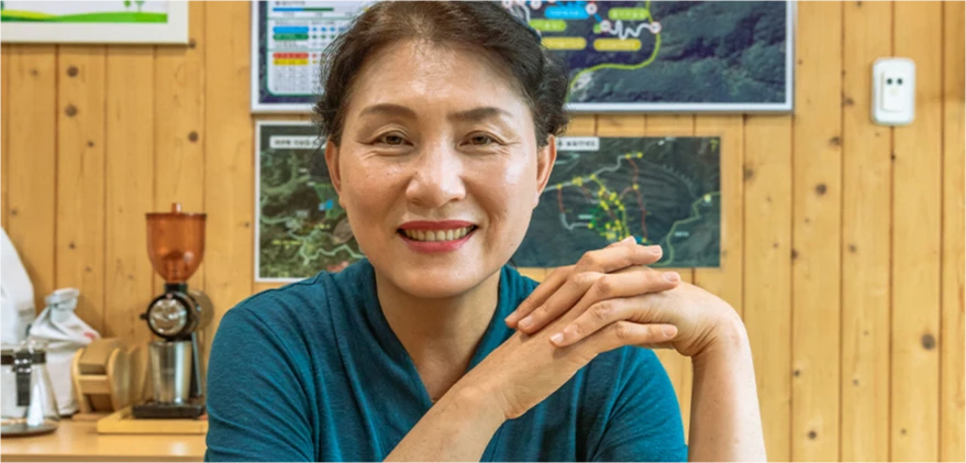 지도 자료가 걸린 실내에서 두 손을 맞잡고 환하게 미소 짓는 치유의 숲 센터장