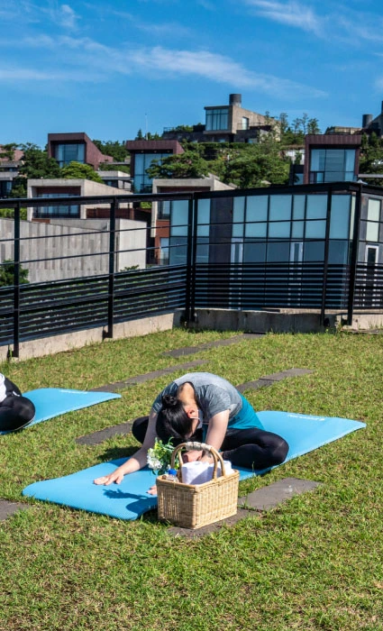 두 참가자가 매트에 앉아 두 팔을 머리 위로 뻗으며 척추를 늘리는 요가 동작을 하는 뒷모습