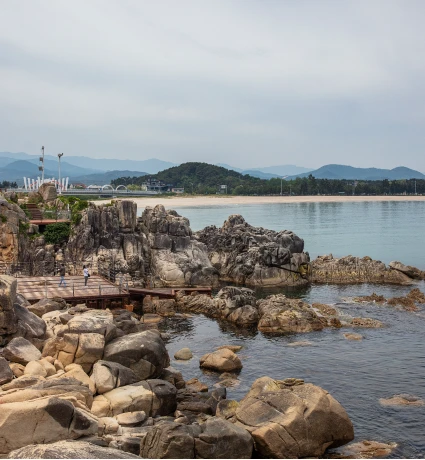 잔잔한 바다 건너 숲이 우거진 곶에 흰 해수관음상이 서 있고 백사장과 마을이 이어진 원경