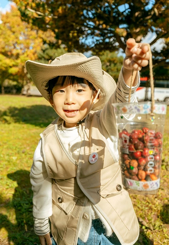 모자와 베스트를 갖춘 남자아이가 빨간 열매가 가득 담긴 채집컵을 줄에 매달아 들어 보이는 모습
