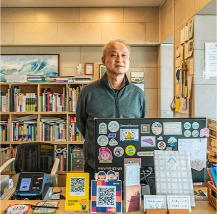 스티커가 가득 붙은 안내 보드 뒤 계산대에 선 서점 대표, 뒤로 책장이 늘어선 매장