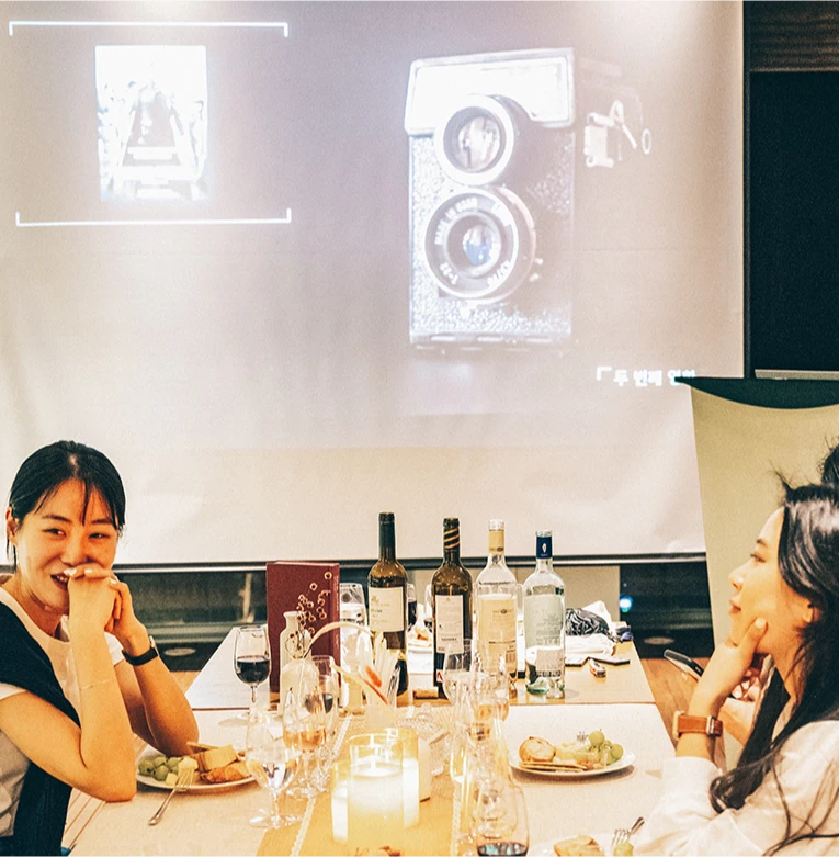 옛 카메라 영상이 비치는 스크린 아래 와인 병과 안주가 놓인 테이블에 마주 앉아 웃는 참가자들