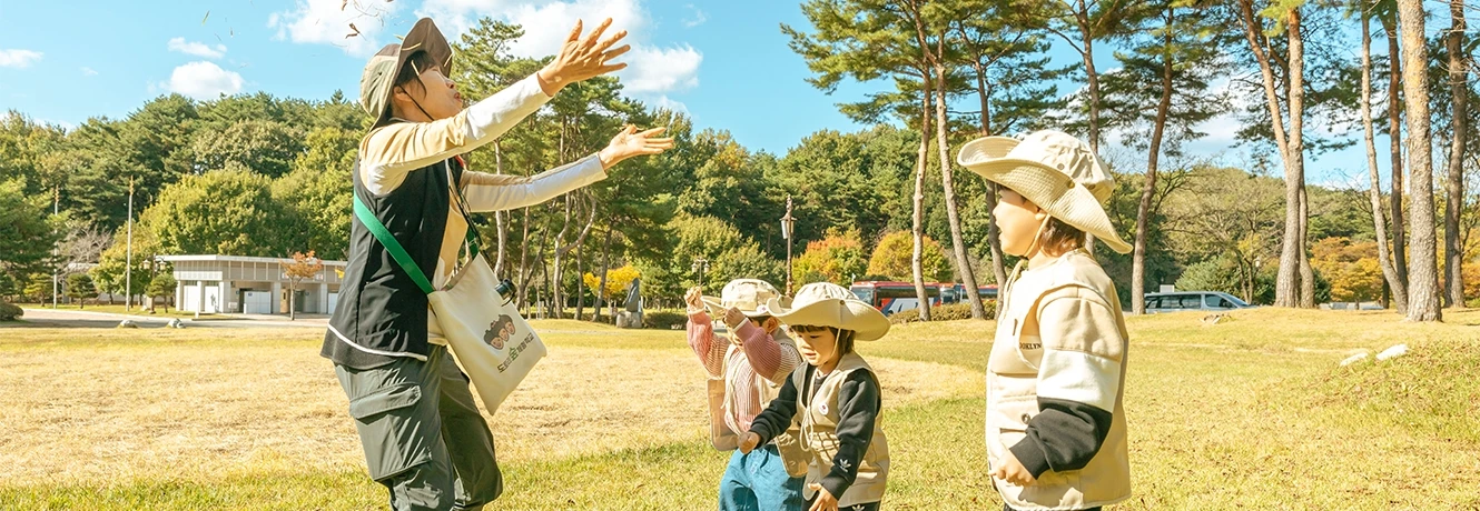 해설사 선생님이 잔디밭에서 칡공을 던지자 모자 쓴 아이들이 둘러서 지켜보는 모습