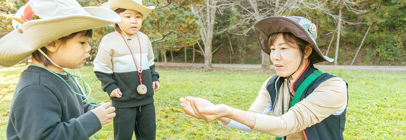 해설사 선생님이 잔디밭에서 두 아이에게 곤충 피리를 들어 보이며 설명하는 모습