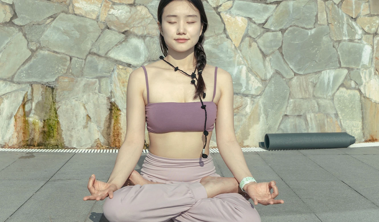돌담을 배경으로 보라색 상의를 입은 여성이 가부좌를 틀고 눈을 감은 채 평온하게 명상하고 있다