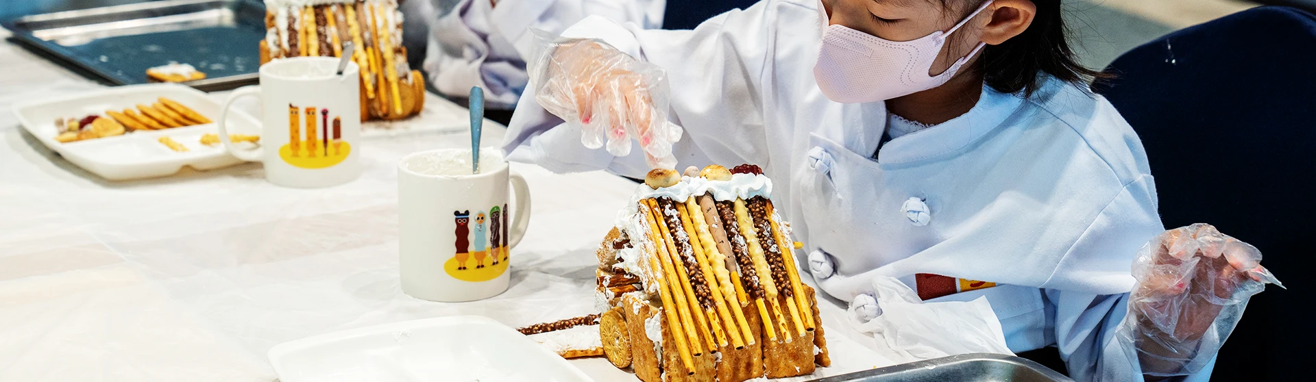 요리사 복장을 한 아이가 빼빼로 과자로 쌓아 만든 과자집을 손으로 꾸미고 있는 모습