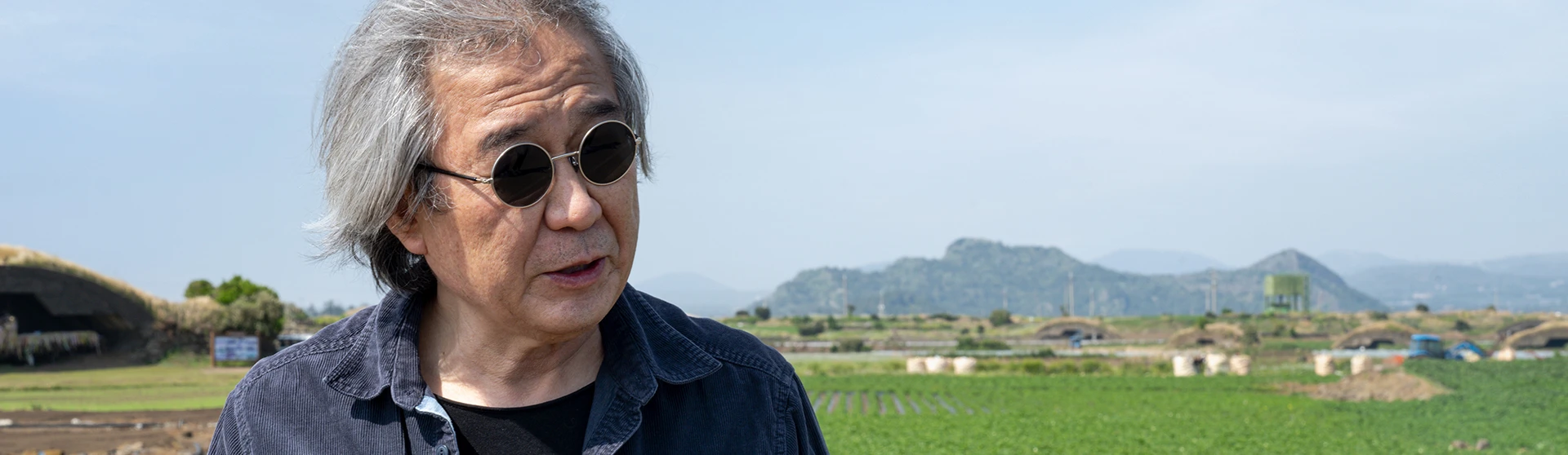 백발에 둥근 선글라스, 네이비 셔츠 차림의 노년 건축가가 산방산과 들판을 배경으로 서 있는 모습