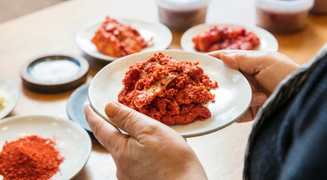 손질된 양념 재료 그릇들이 놓인 식탁에서 한 손으로 가자미식해가 담긴 흰 접시를 들어 보이는 모습