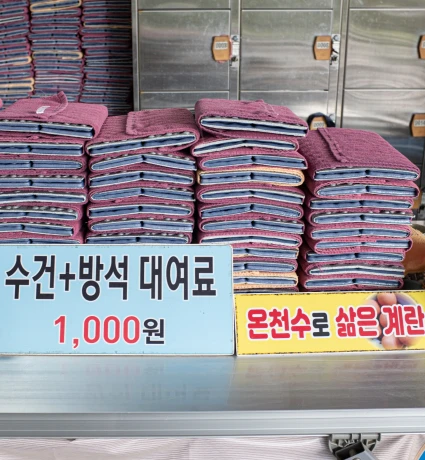라커 앞 매대에 수건과 방석이 쌓여 있고 대여료 1,000원과 온천수 삶은 계란 안내판이 놓인 모습