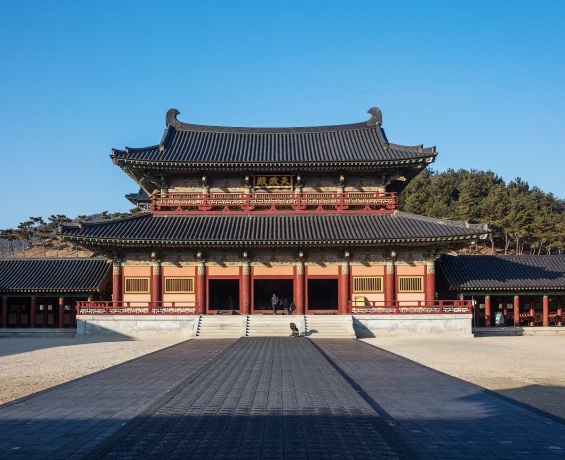 붉은 기둥과 큰 기와지붕을 갖춘 백제문화단지 천정전 본전이 너른 돌마당 너머 푸른 하늘 아래 자리하고 있다