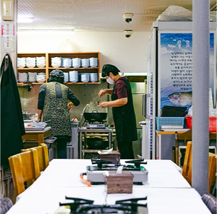 흰 식탁이 놓인 식당 안쪽 주방에서 직원들이 물곰탕을 끓이며 음식을 준비하는 사돈집 내부
